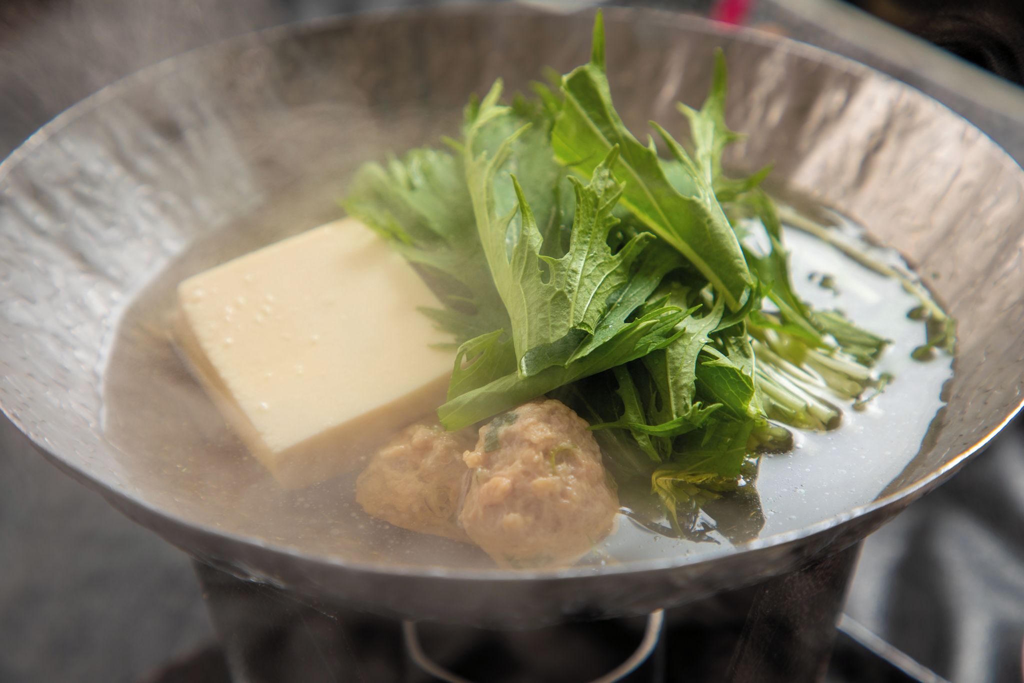A hot pot with tofu, meatballs, and greens in a metal pot.