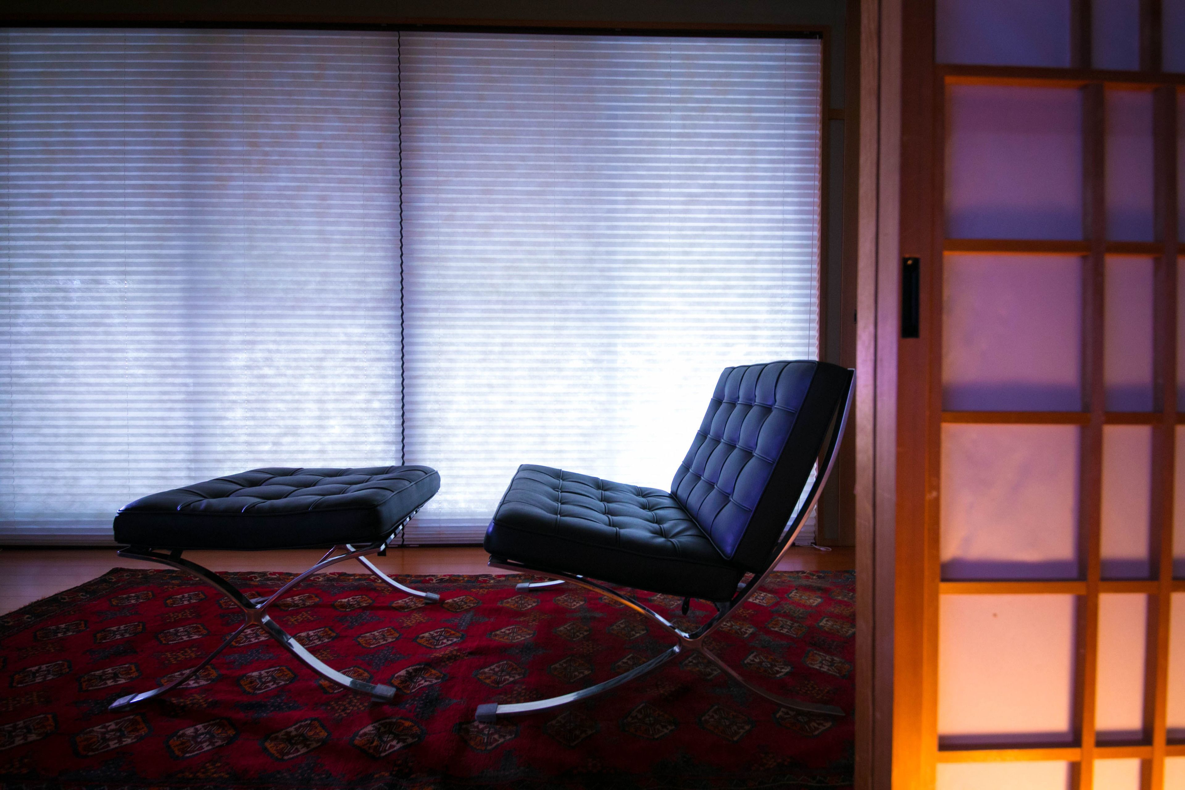 A room with a black leather chair and ottoman on a patterned rug, a sliding glass door, and a view through the blinds.
