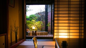 Una habitación japonesa tradicional con suelo de tatami, una mesa baja, una silla y vistas a un jardín a través de una puerta corredera.