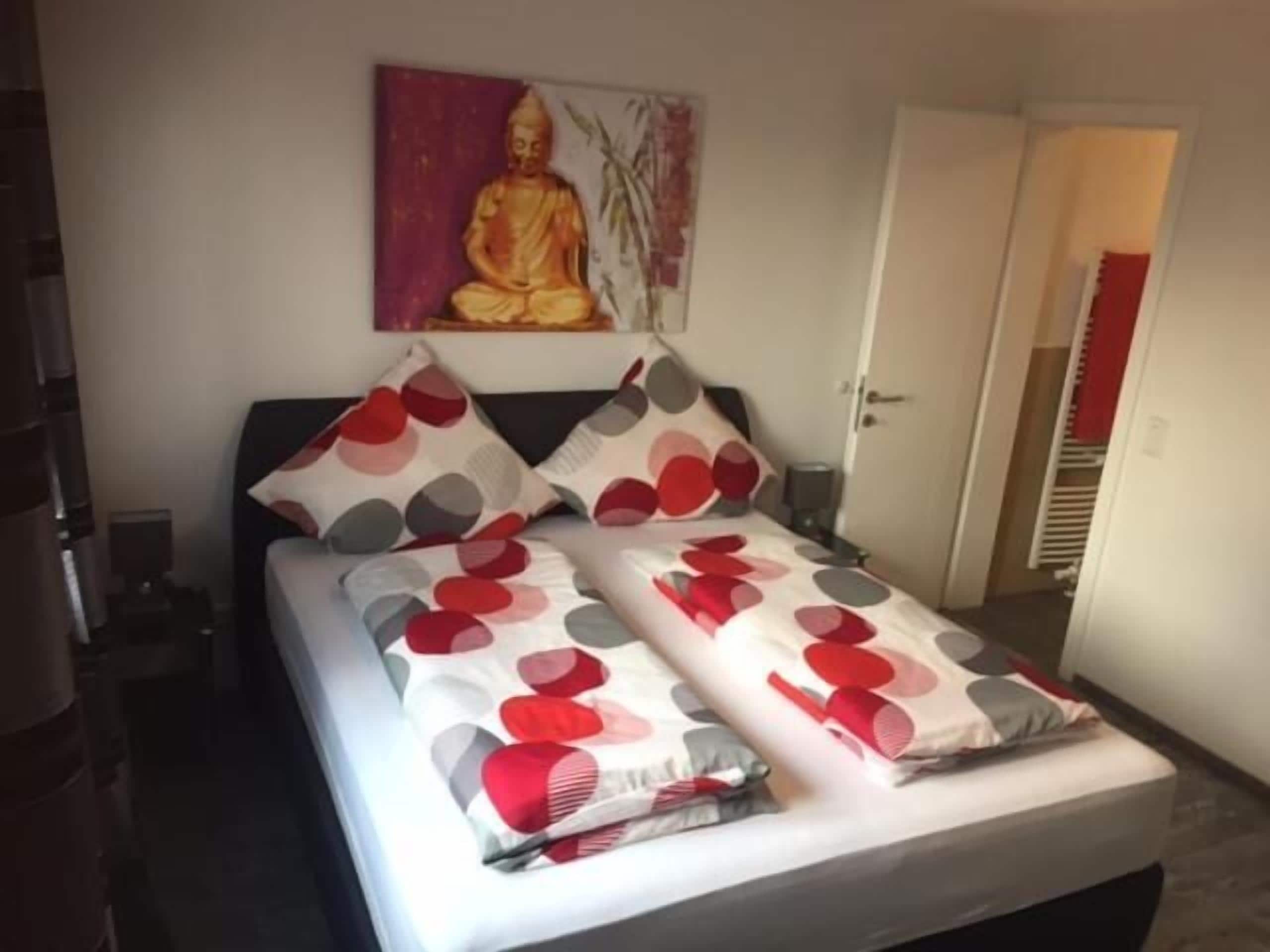 A neatly made bed with patterned pillows, a wall painting, and a door leading to another room.