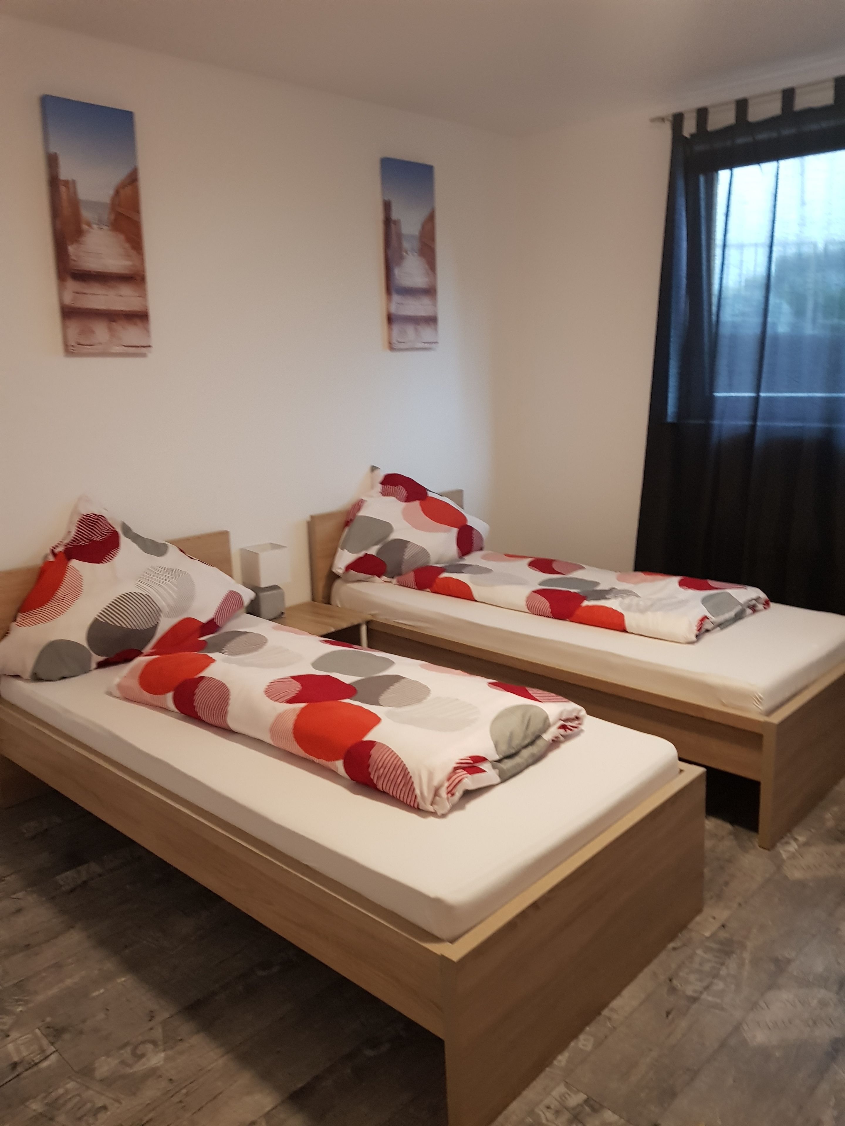 Quarto de hotel com duas camas de solteiro, cada uma com colcha e almofadas estampadas. Há duas obras de arte na parede retratando paisagens. O quarto tem piso de madeira e uma janela grande com cortinas escuras.