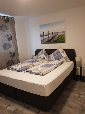 Una cama bien hecha con una colcha estampada y almohadas, un cuadro de un muelle en la pared y un suelo de madera.