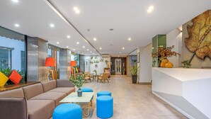 A modern hotel lobby with a sectional sofa, blue ottomans, a reception desk, and a large leaf artwork on the wall. - Platinum Danang Hotel (Da Nang)