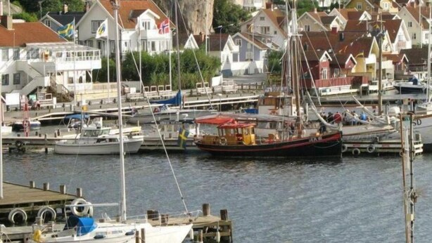 A marina with boats docked, a small boat with a red canopy, and houses with flags in the background. - Fjällbacka Pensionat & Vandrarhem (Fjallbacka)