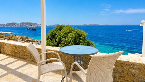 A terrace with a table and chairs overlooking a clear blue sea, with a ship in the distance. - Villa Oceania (Mykonos)
