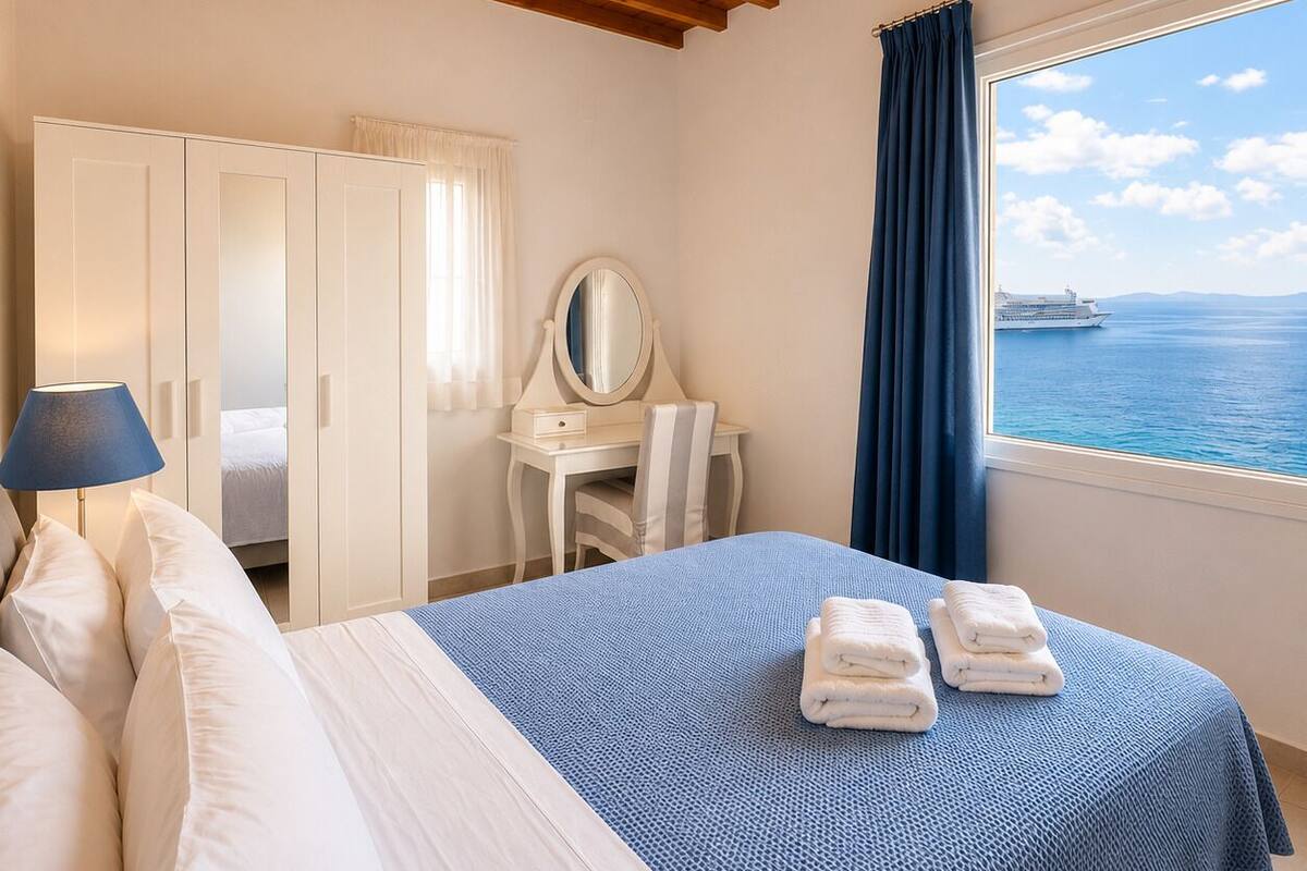 A bedroom with a bed, a desk with a mirror, a wardrobe, and a view of the sea.