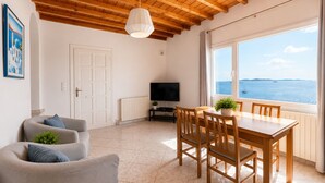 A living room with a wooden ceiling, a television, a dining table with chairs, and a large window with a view of the sea. - Villa Oceania (Mykonos)