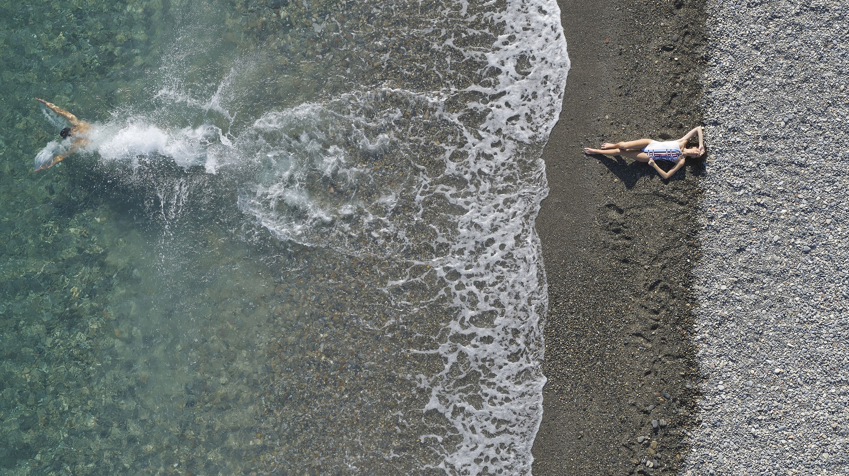 An aerial view of a beach with clear water and a person swimming. Another person is lying on the sand near the water's edge.