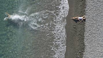 An aerial view of a beach with clear water and a person swimming. Another person is lying on the sand near the water's edge.
