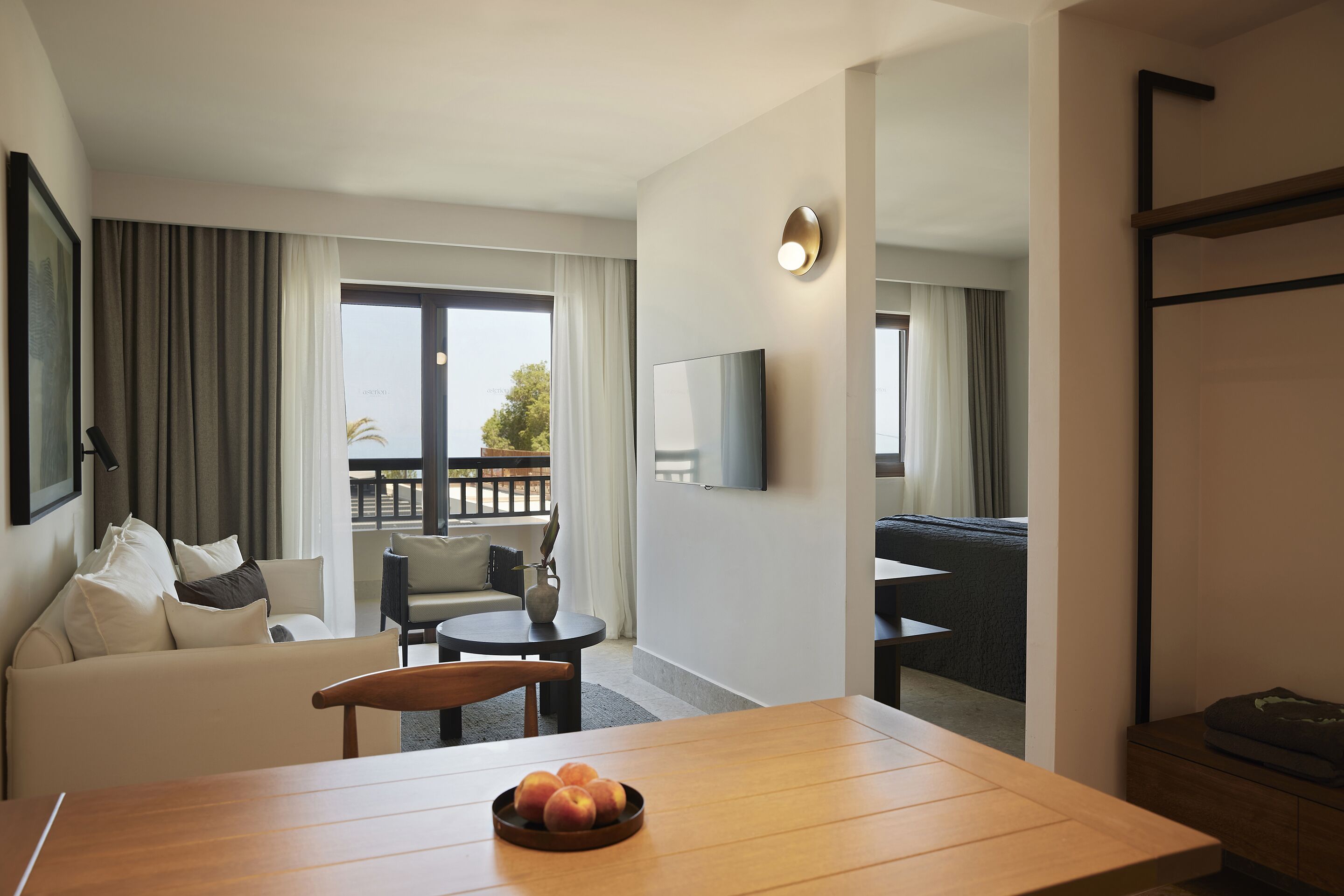 A modern hotel room with a wooden dining table, a sofa, a TV mounted on the wall, and a balcony with a view.