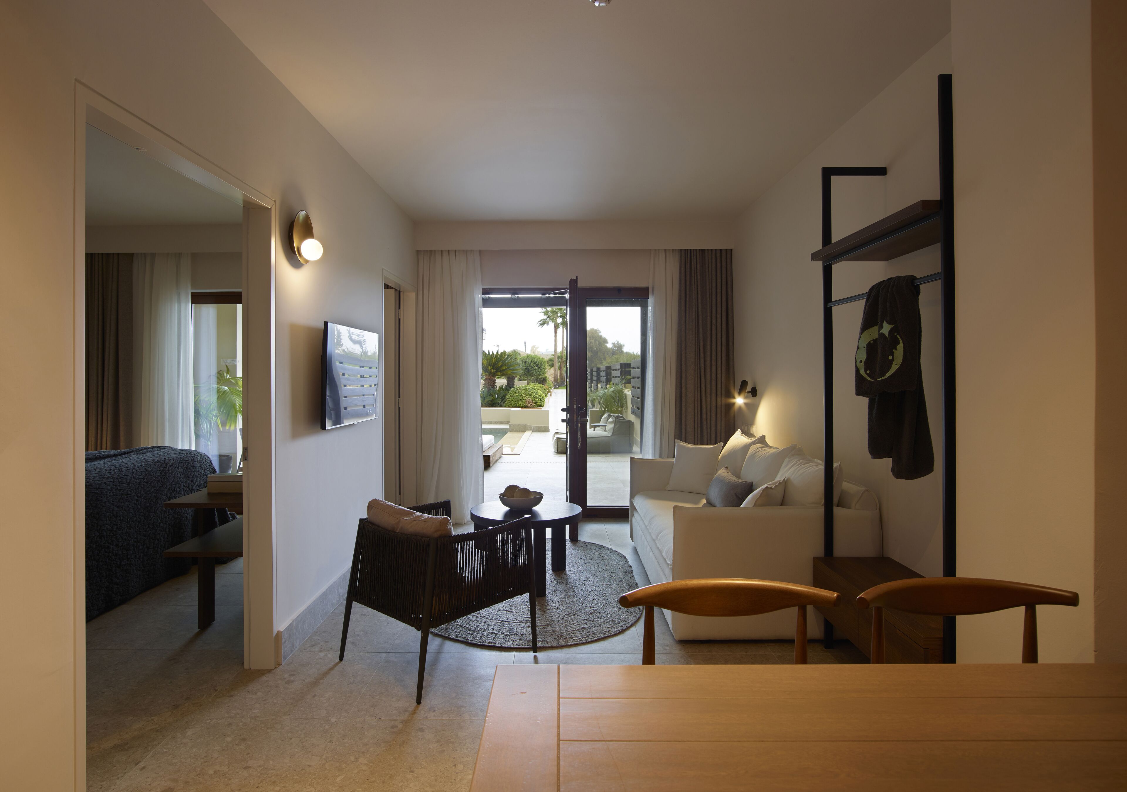 A modern hotel room with a sofa, a dining table, a chair, a TV, and a view of the outdoor area.