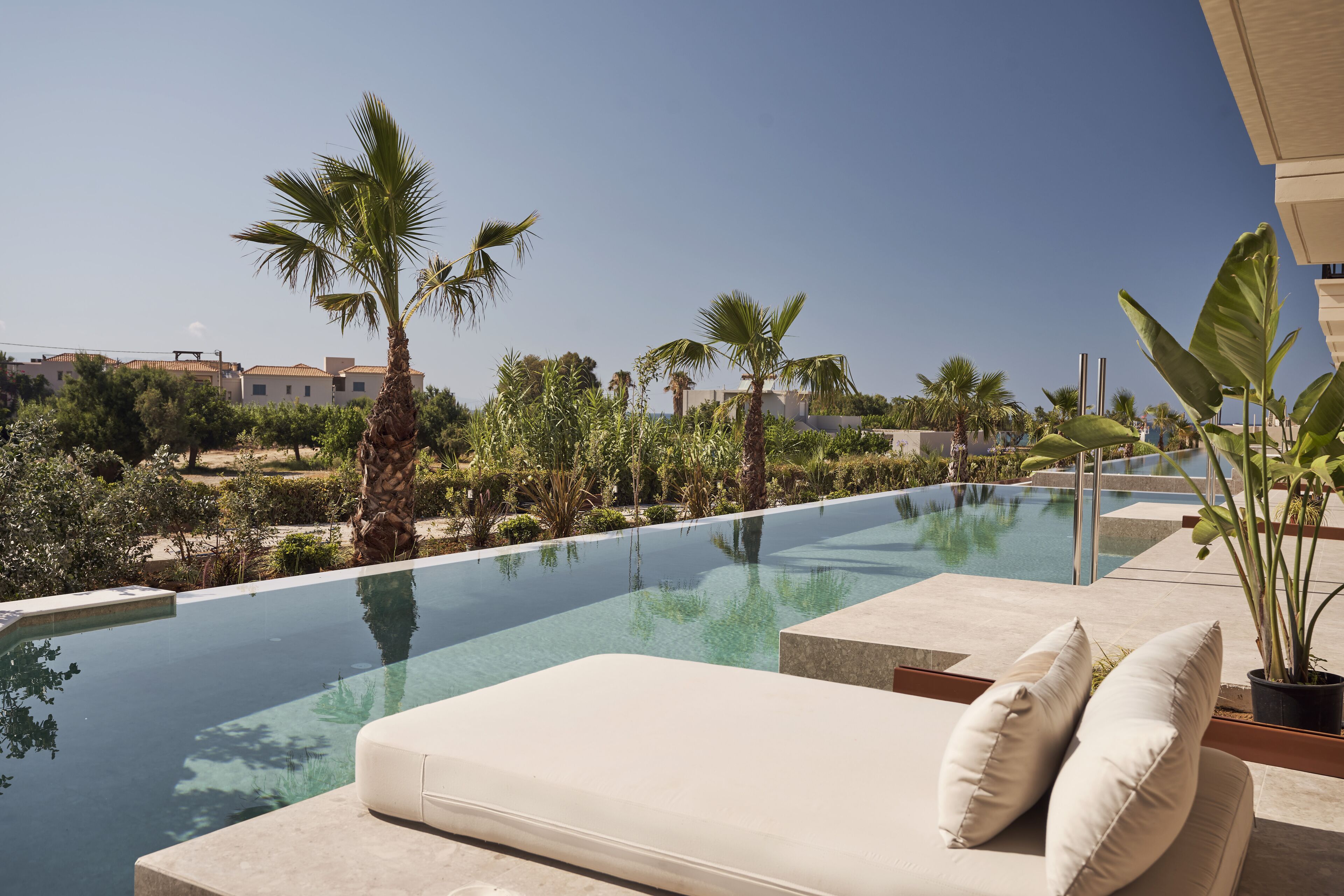 Una piscina infinita con una silla de descanso blanca y cojines, rodeada de palmeras y follaje.