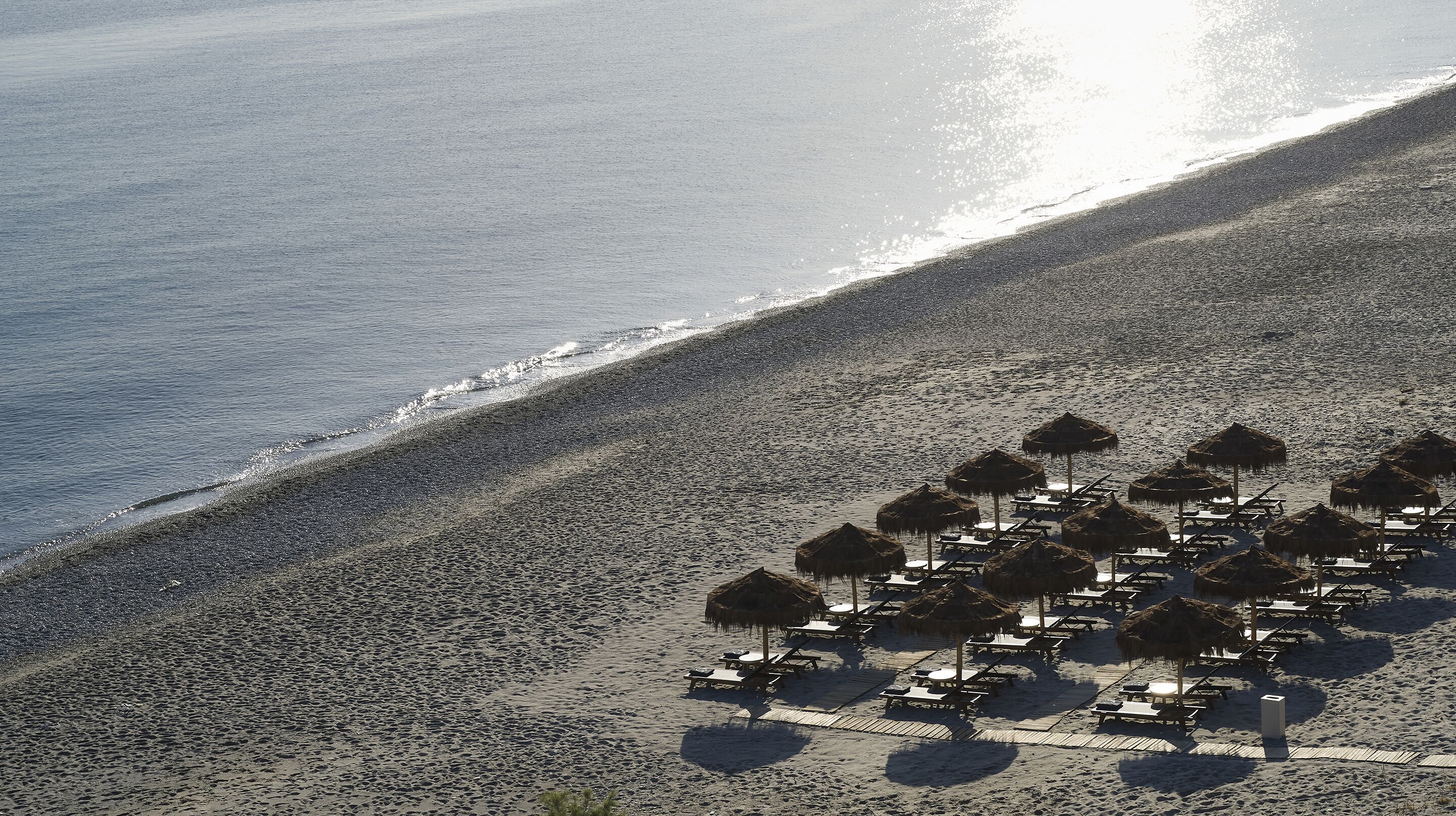 An aerial view of a beach with thatched umbrellas and sun loungers arranged along the shoreline.