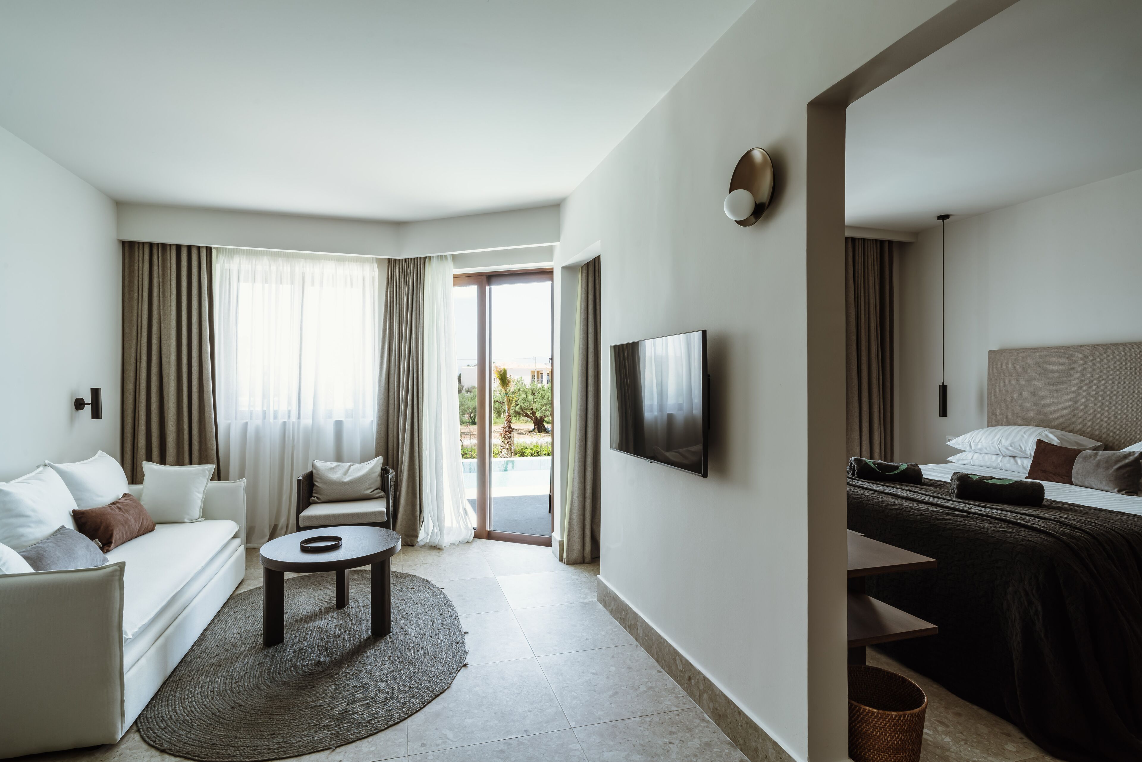 Una habitación de hotel moderna con sofá, una mesita redonda, cama con cabecera y televisión en la pared.