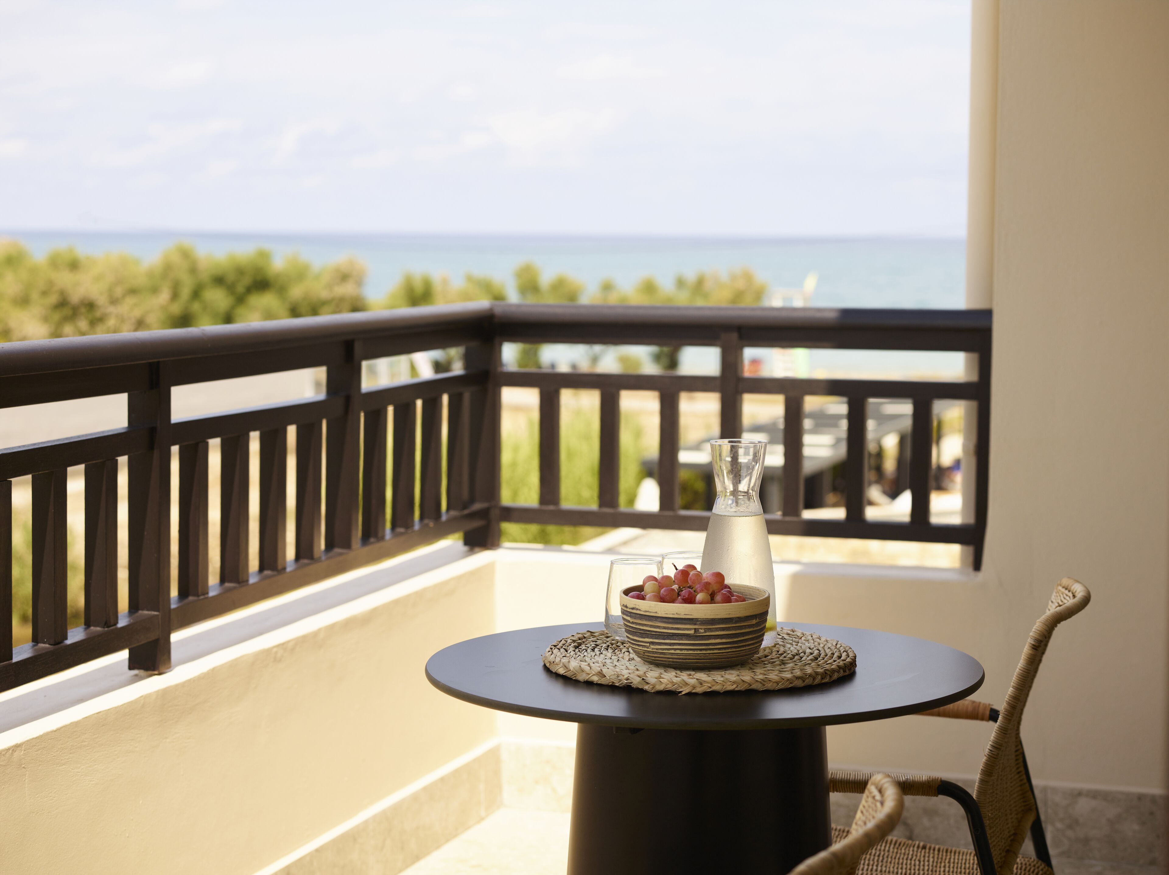 A balcony with a round table, a wicker chair, a glass pitcher, and a bowl of grapes.