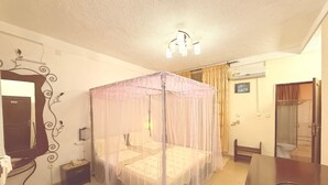 A bedroom with a four-poster bed, a bedside table, a mirror, a small bedside table, a chair, and a bathroom with a shower and toilet. - Gahahe Hills Hotel (Bujumbura)