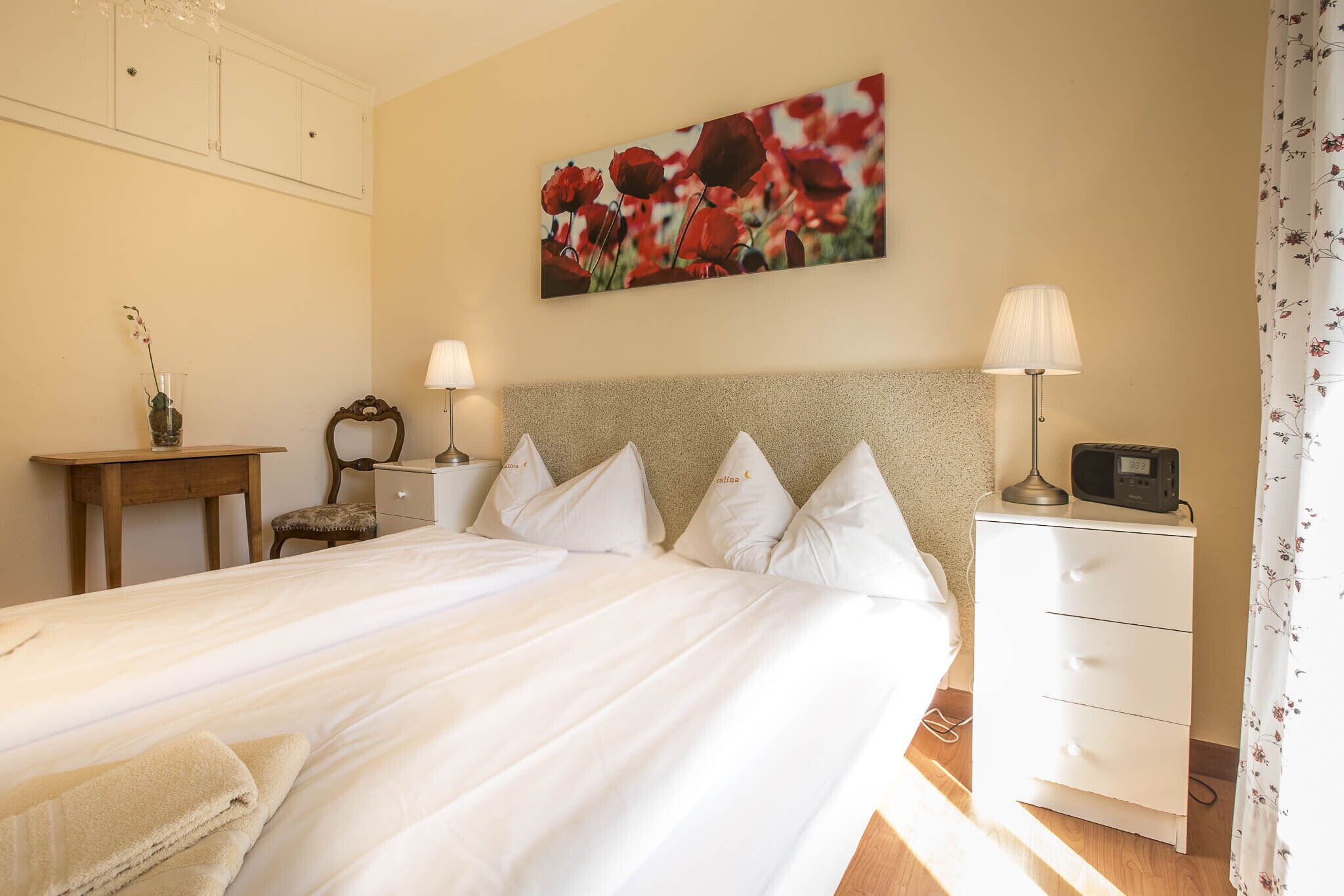 Un lit bien fait, avec du linge de lit blanc, une table de chevet avec une lampe, une petite table de nuit avec une radio et un tableau floral accroché au mur.