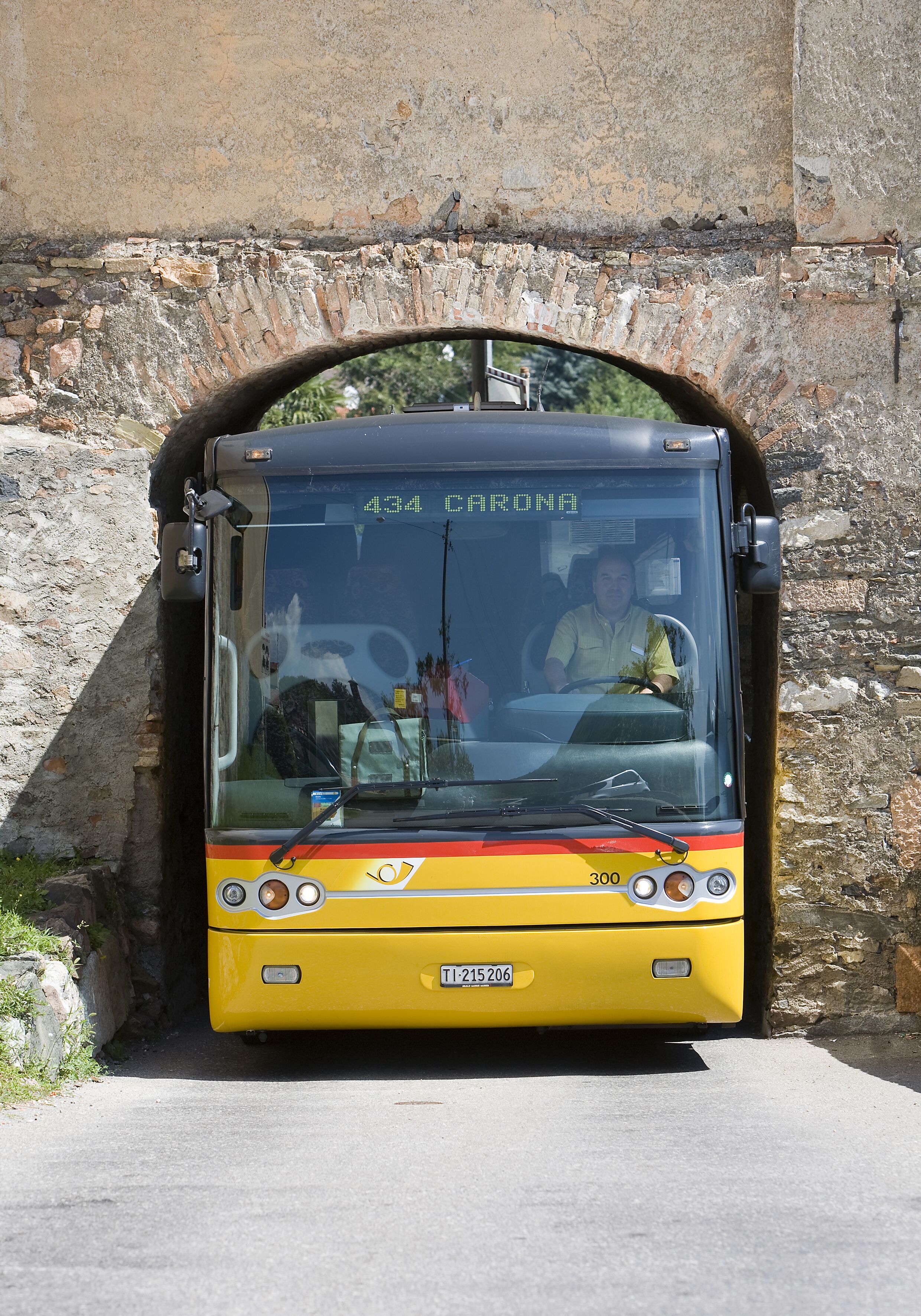 Ein gelber Bus mit der Nummer 434 und dem Zielort 'CARONA' fährt durch ein steinernes Tor.
