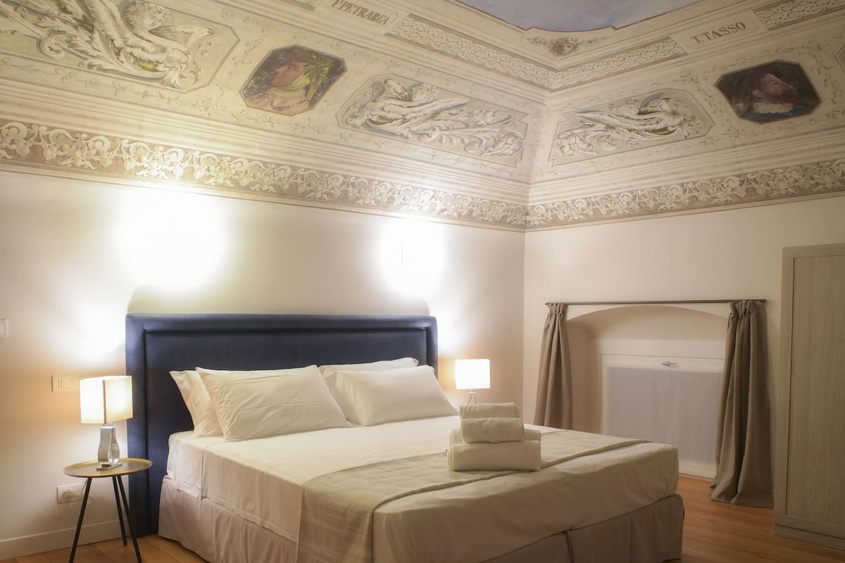 Una habitació d'hotel amb un llit gran, una meseta de nit amb una làmpada i un sostre amb treballs artístics detallats.
