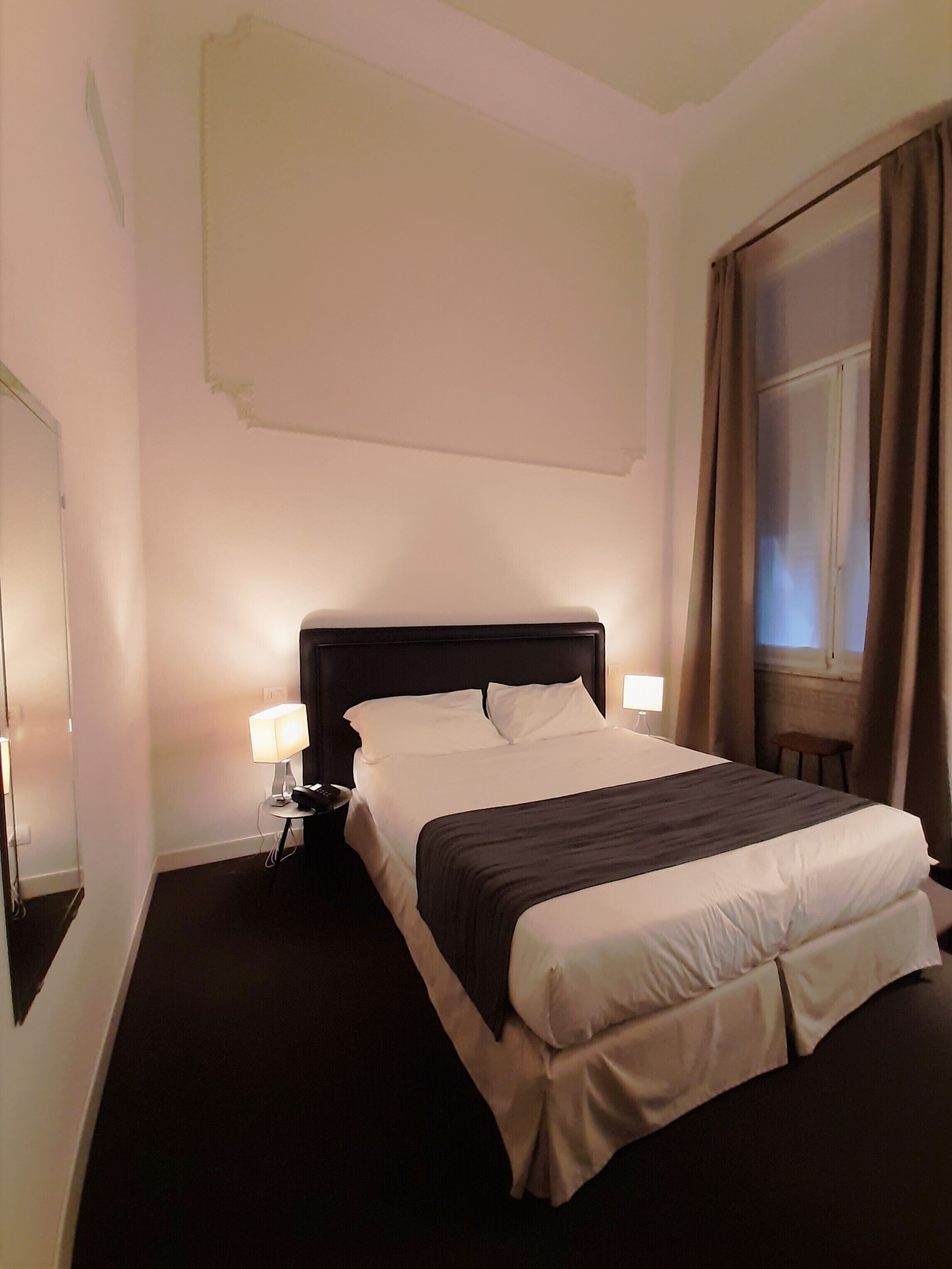 A hotel room with a large bed, two bedside tables with lamps, a window with curtains, and a plain white wall.