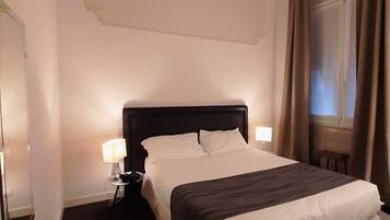 Une chambre d’hôtel avec un grand lit, deux tables de chevet avec des lampes, une fenêtre avec des rideaux et un mur blanc uni.