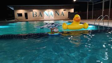Une piscine avec des jouets gonflables, un dauphin et un canard. En arrière-plan, on aperçoit un bâtiment sur lequel le mot « BANANA » est illuminé.