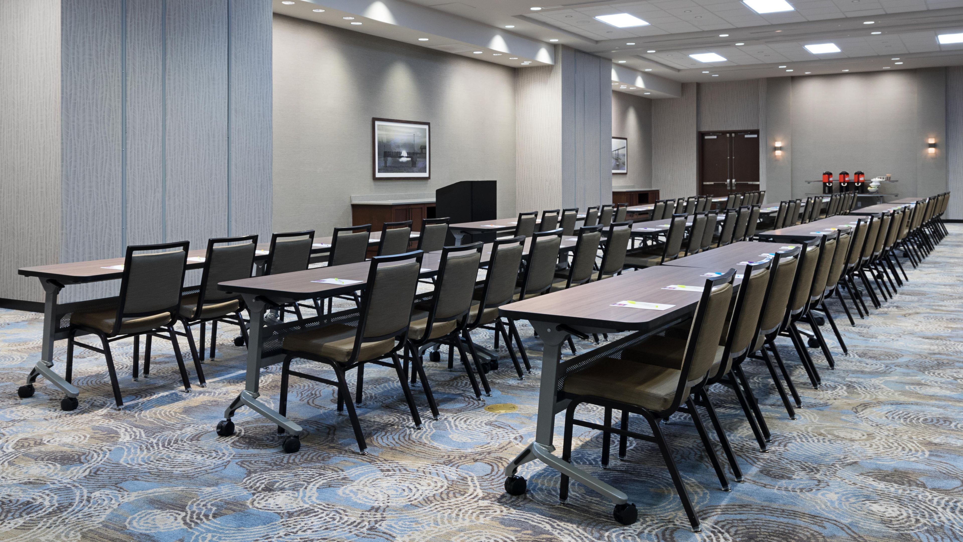 Un salón de conferencias con filas de mesas y sillas, un atril y piso alfombrado.