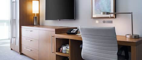 A hotel room with a wooden desk, a chair, a flat-screen TV, a lamp, a telephone, and a shelf with toiletries.