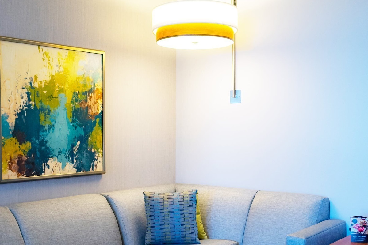 A modern living room with a sectional sofa, a small ottoman, a wall-mounted lamp, and a colorful abstract painting.