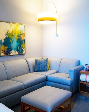 A modern living room with a sectional sofa, a small ottoman, a wall-mounted lamp, and a colorful abstract painting. - Hyatt Place St. Petersburg / Downtown (St. Petersburg)