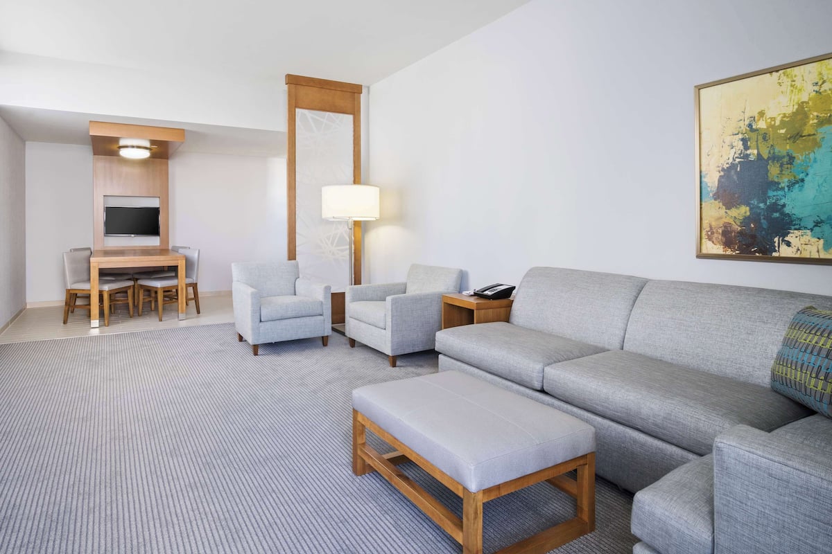 A modern hotel room with a grey sofa, two armchairs, a wooden coffee table, a TV, a desk with a chair, and a floor lamp.