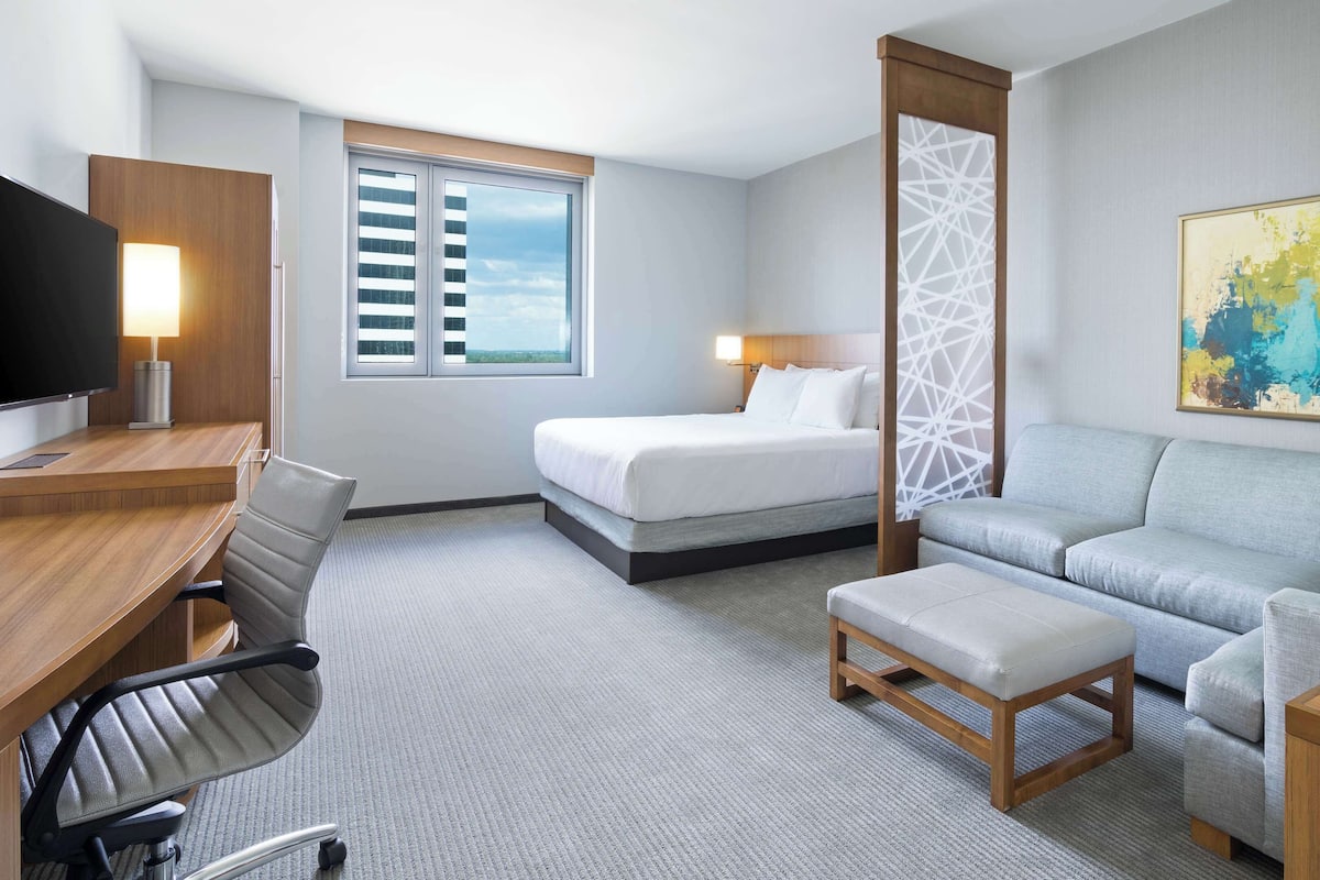 A hotel room with a bed, a desk with a chair, a sofa, a small ottoman, a TV, and a painting on the wall.