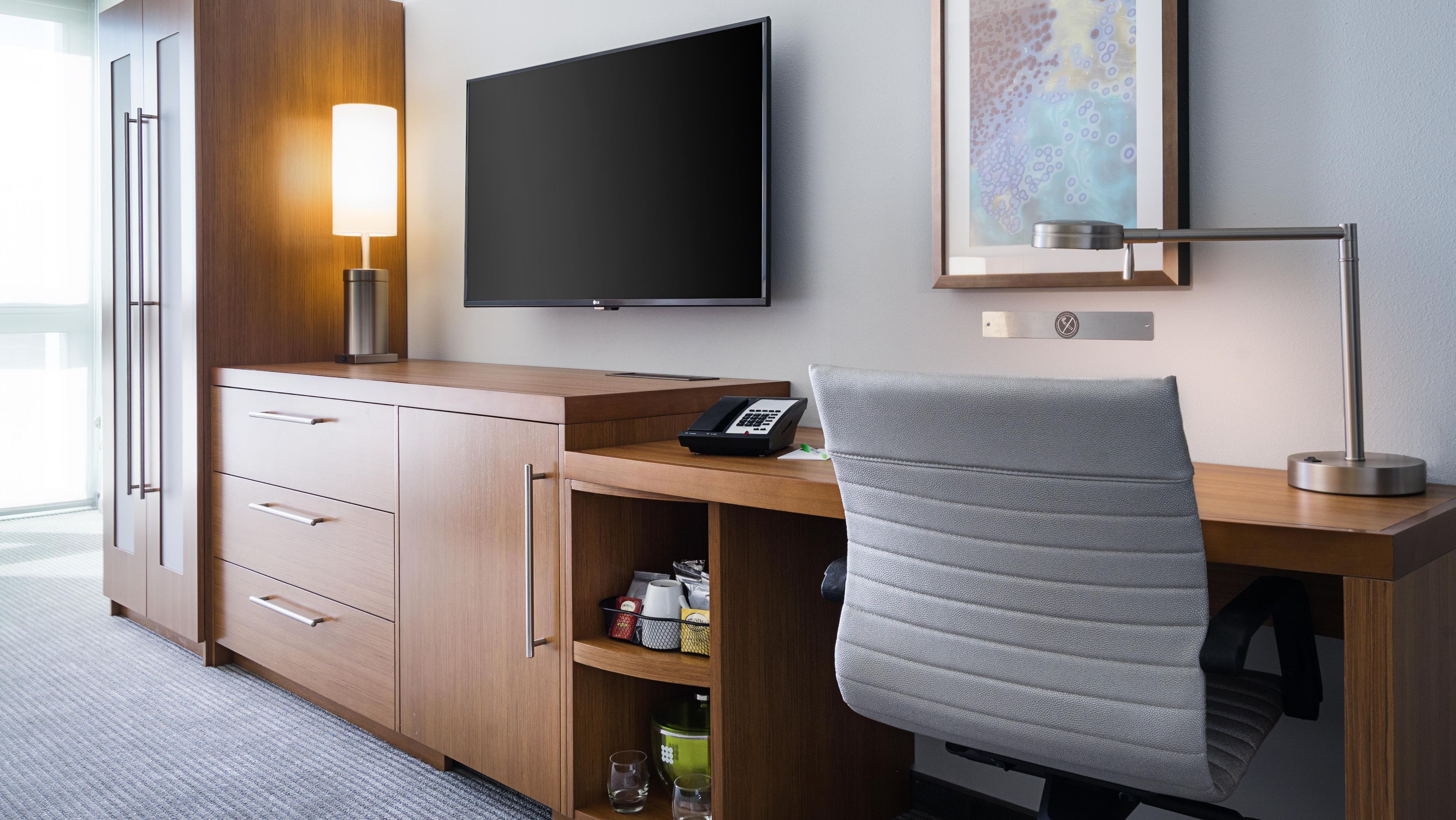 Quarto de hotel com escrivaninha, cadeira, televisão e prateleira com objetos.