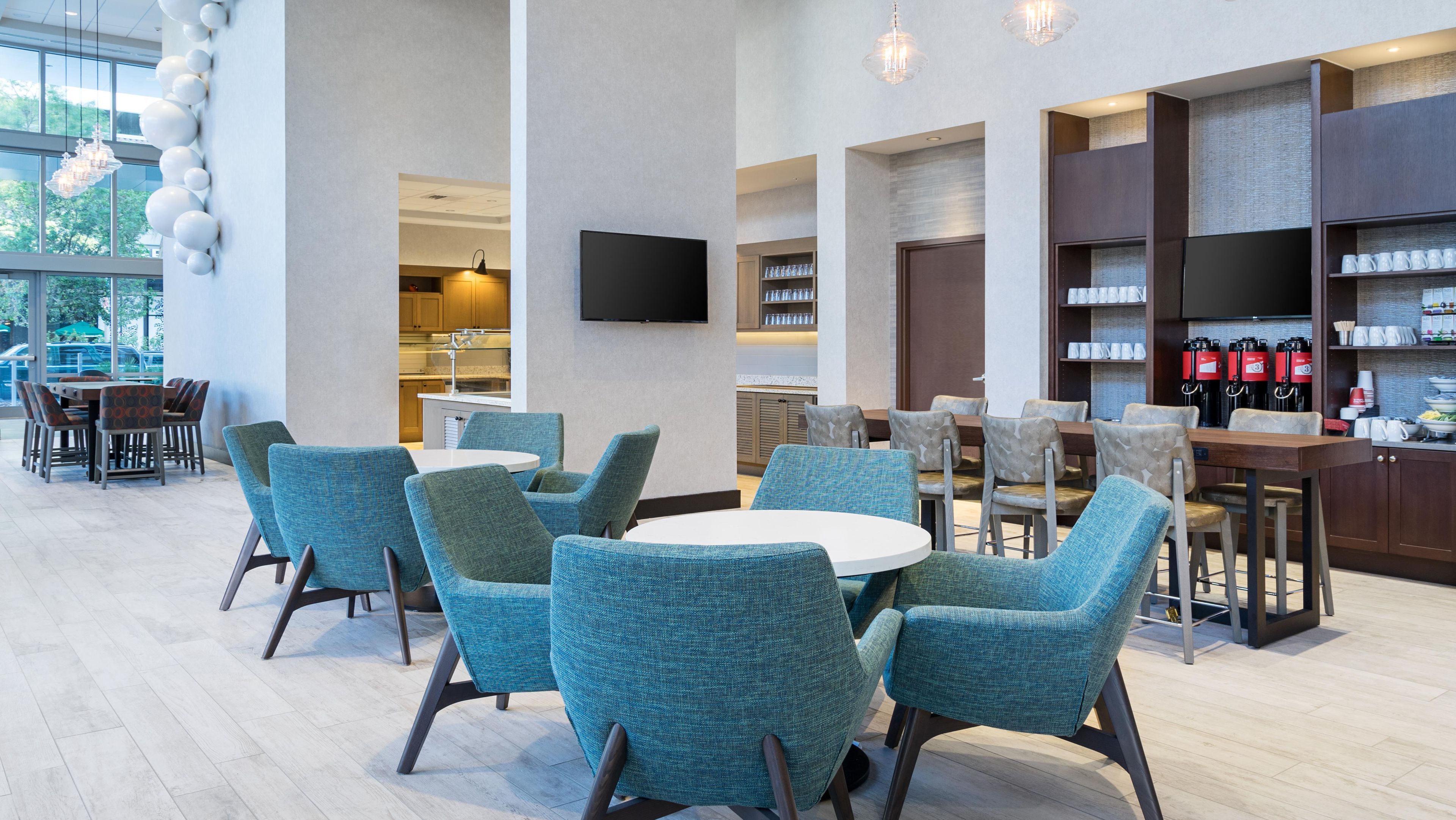 A modern dining area with round tables and teal chairs, a kitchen area with a flat-screen TV, and a bar with coffee machines.