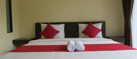A neatly made bed with white and red bedding, two red pillows, and a pair of rolled towels on the bed.