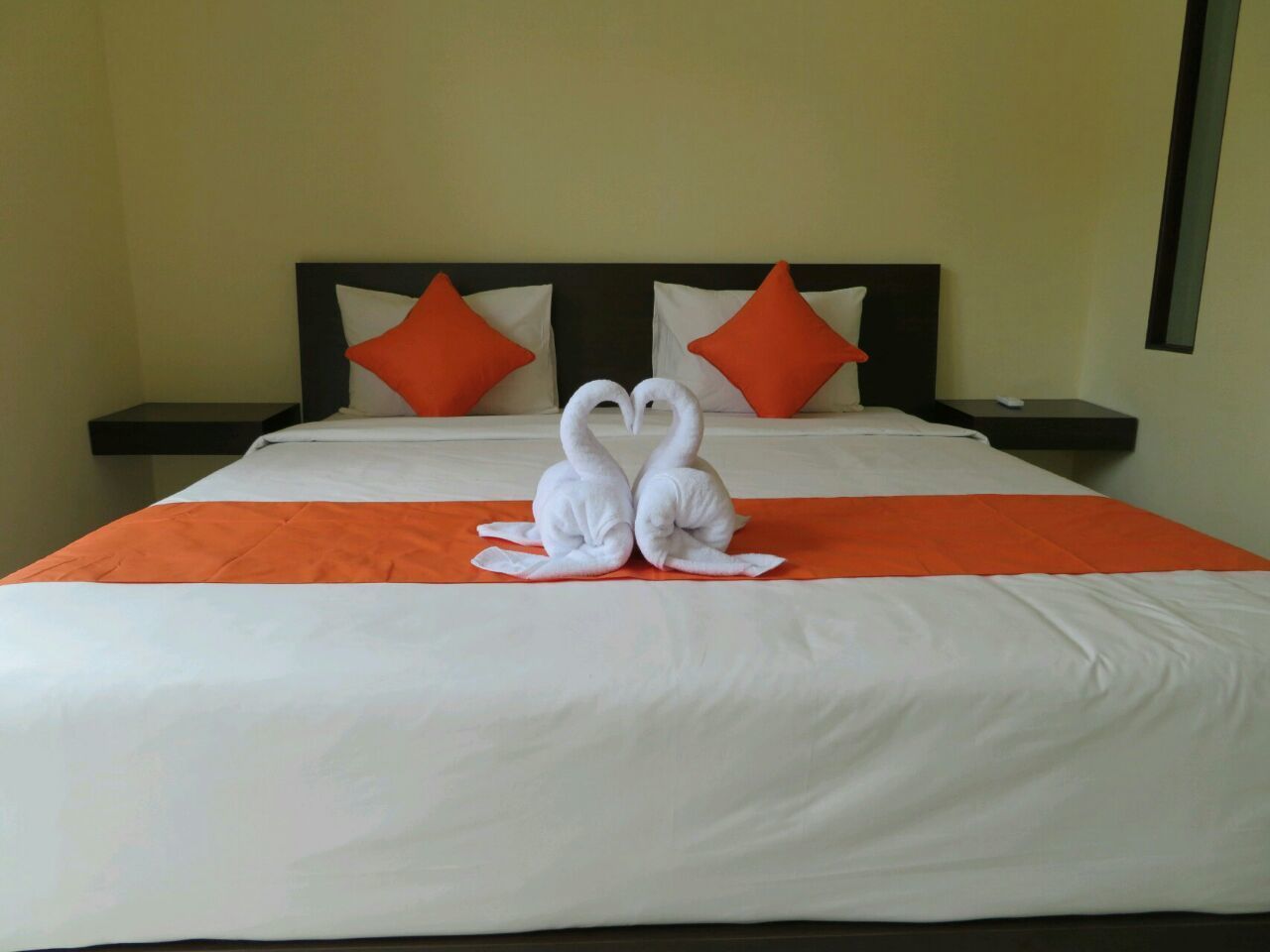 A hotel room with a bed, orange and white bedding, two orange pillows, and two white towels arranged in a heart shape on the bed.