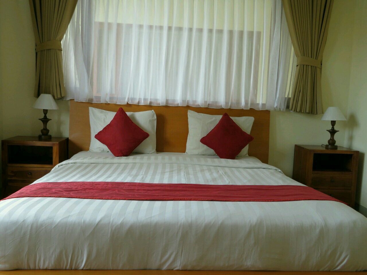 A neatly made bed with a red throw blanket and two red pillows, flanked by two bedside tables with lamps, in a room with sheer curtains and a wooden headboard.