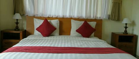 A neatly made bed with a red throw blanket and two red pillows, flanked by two bedside tables with lamps, in a room with sheer curtains and a wooden headboard.