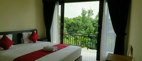 A hotel room with a large bed, a small table, and a balcony with a view of greenery.