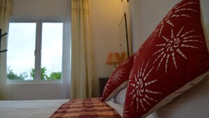 A bed with a checkered blanket, a red pillow with a sun design, and a window with curtains. - Sunshine View (Maafushi)