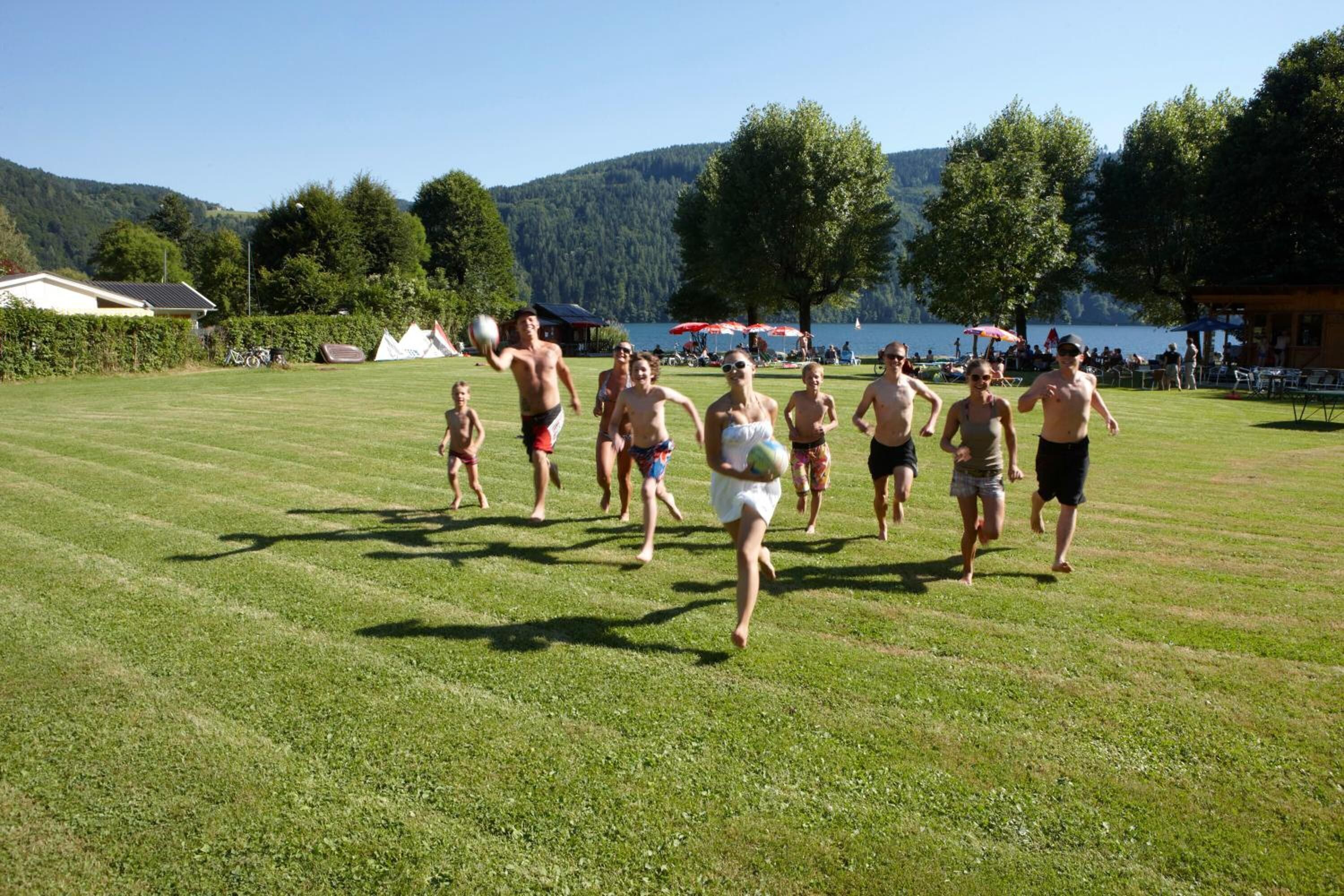 A grassy field with people running, a lake, and trees in the background.