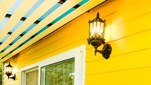 A yellow building with a sliding glass door, a metal shelf with flowers, and a wall-mounted light fixture. - Sweet Lighthouse Homestay (Pak Chong)