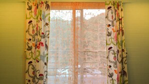 A room with a ceiling fan, a window with patterned curtains, and a view of trees outside. - Sweet Lighthouse Homestay (Pak Chong)