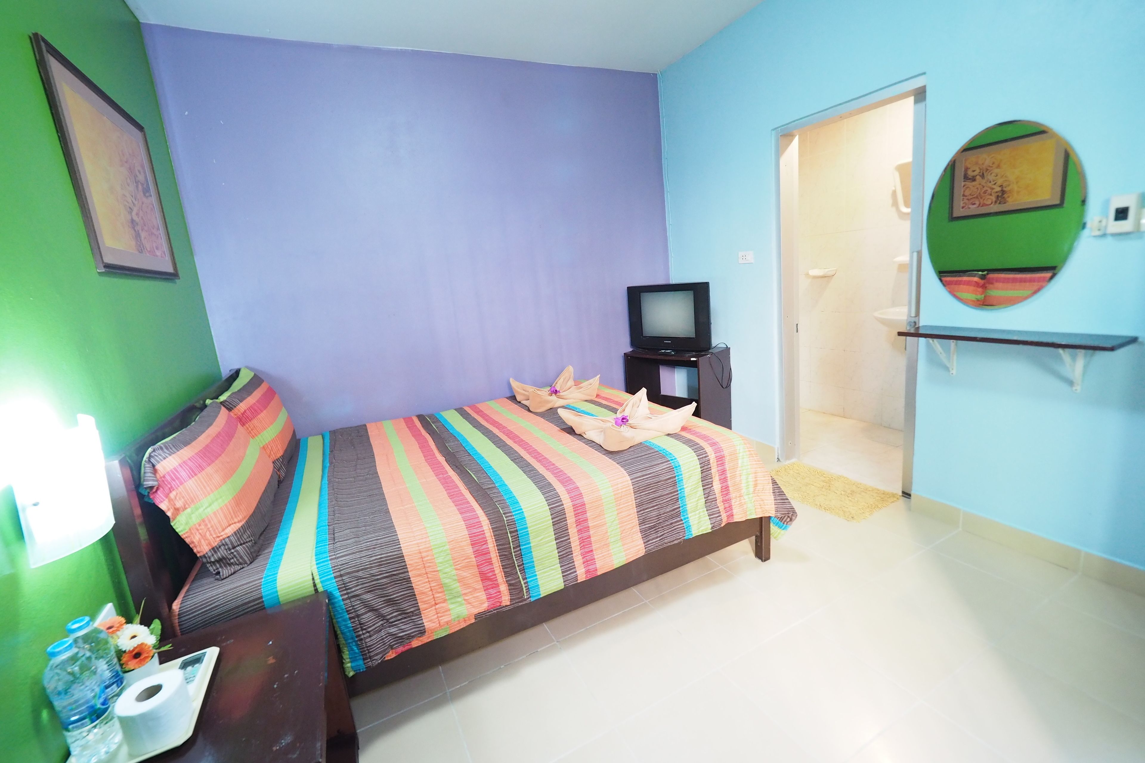A room with a bed, a television, a small table with a bottle and flowers, and a door leading to another room.