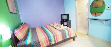 A room with a bed, a television, a small table with a bottle and flowers, and a door leading to another room.