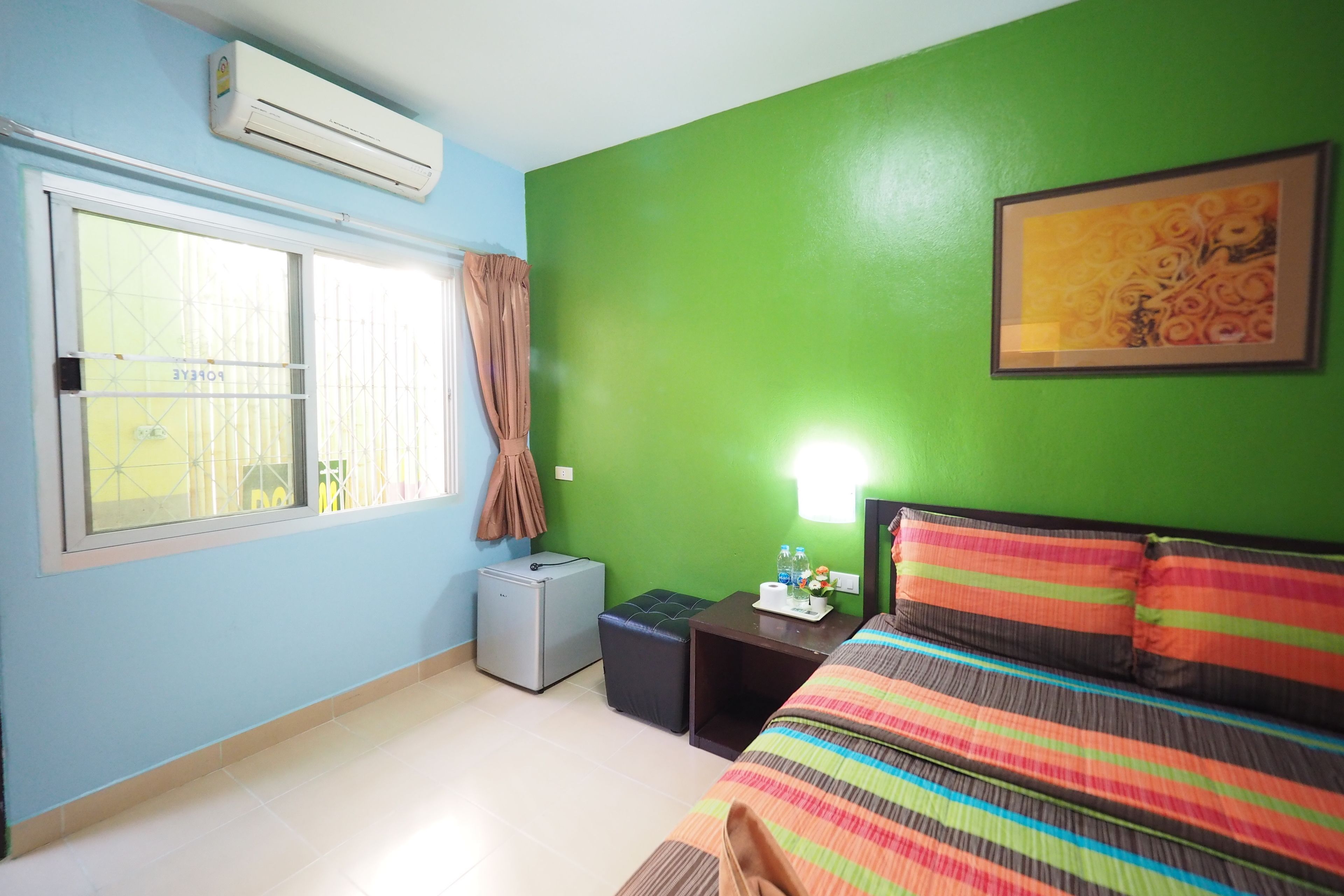 Una habitación de hotel con una pared verde, una cama con ropa de cama a rayas, una mesita de noche con una lámpara y una ventana con cortinas.