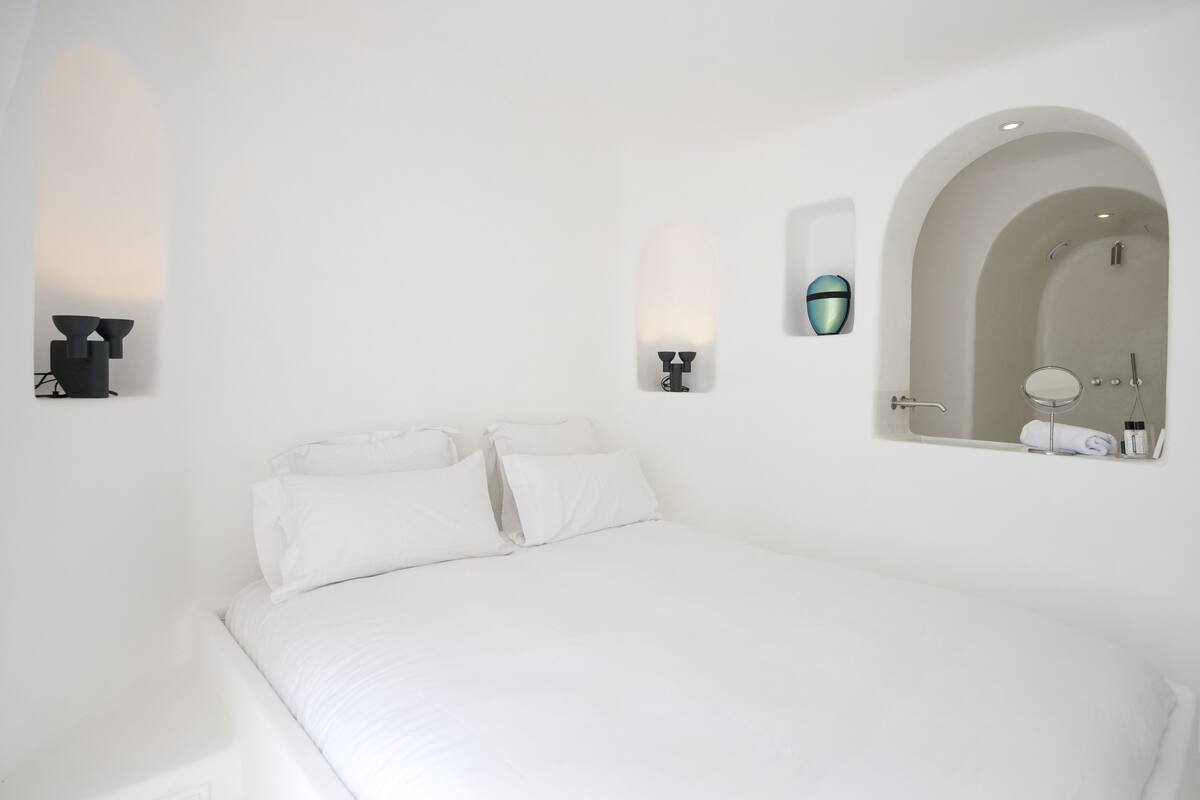 Habitación blanca con una cama, un espejo en la pared y un pequeño estante con artículos de tocador.