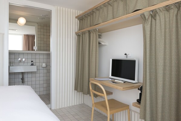 Habitación de hotel con cama, escritorio con computadora, televisor, espejo y baño con lavamanos e inodoro.