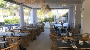 A restaurant with multiple tables and chairs, large windows, and hanging light fixtures. - La Reine Jane (Hyères)