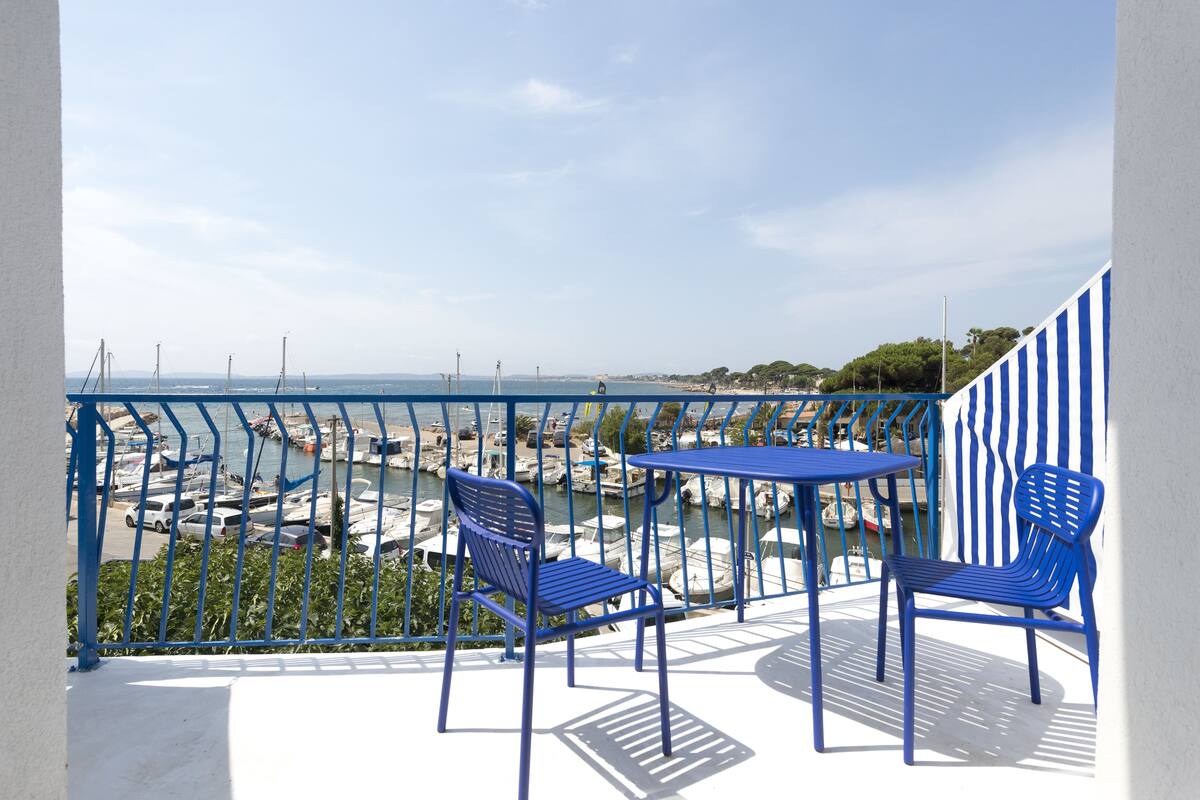 Un balcón con dos sillas azules y una mesa, con vista a un puerto deportivo con barcos y un cielo despejado.